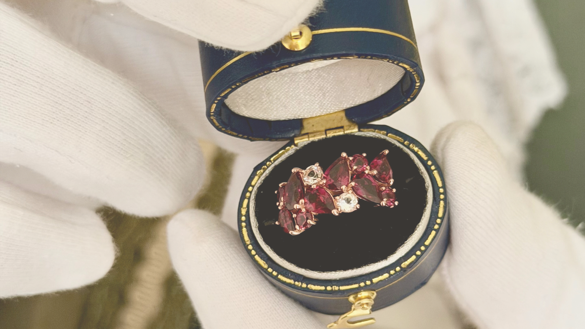 A close-up of a pair of red gemstone rings displayed in an open dark blue and gold ring box, held by white-gloved hands.