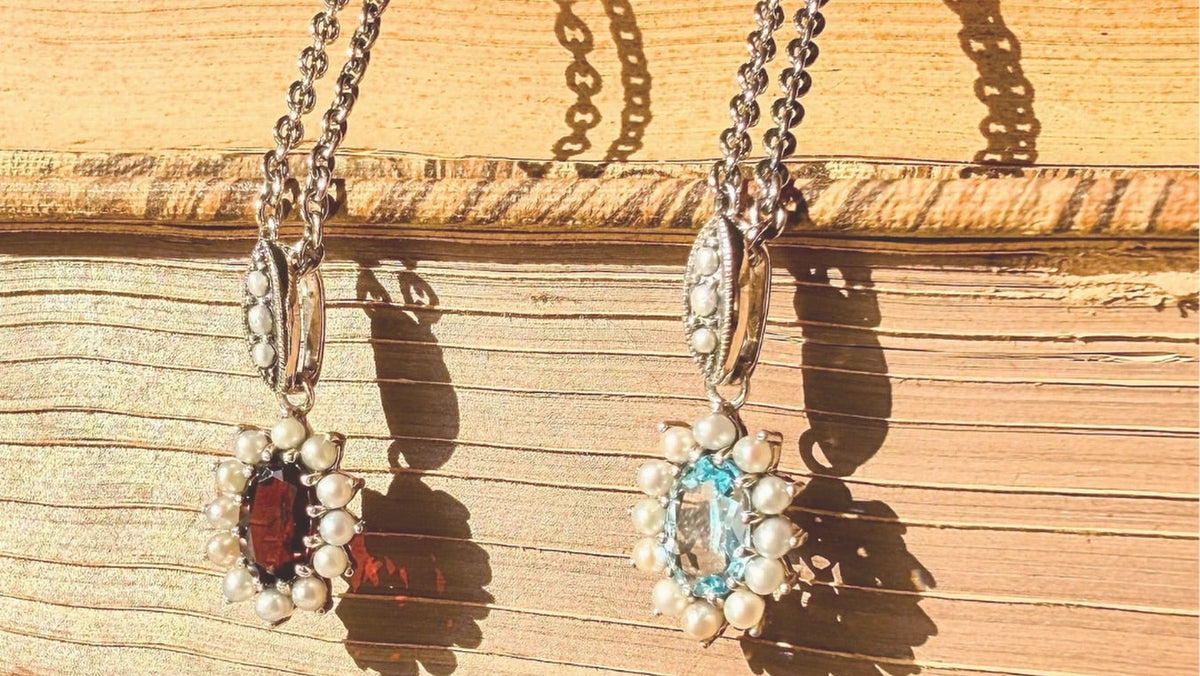 Two silver-toned floral pendant necklaces with pearl-like accents hang against a rustic wooden background. One pendant features a dark red oval stone, and the other features a light blue oval stone.