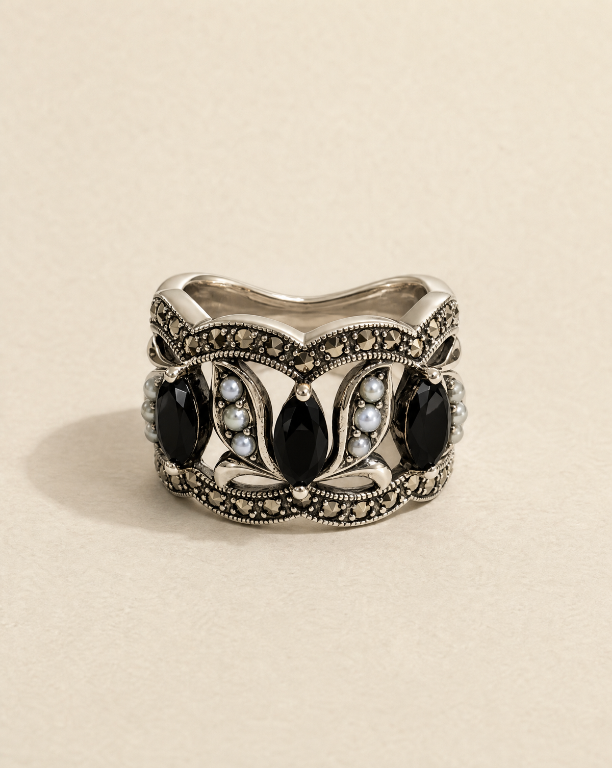 An ornate silver ring featuring five large black teardrop-shaped stones and small pale blue stones, set against a warm beige background.