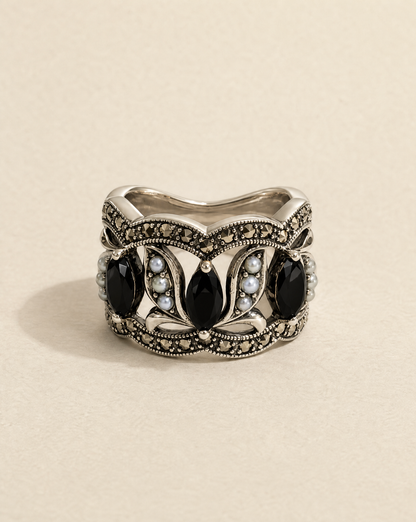 An ornate silver ring featuring five large black teardrop-shaped stones and small pale blue stones, set against a warm beige background.