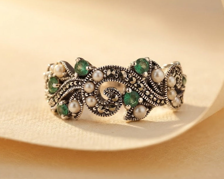 An ornate silver ring with green gemstones and white pearl accents, displayed on a soft beige fabric background.