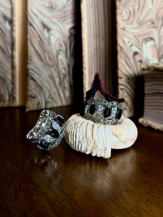 Two ornate silver rings with black gemstones displayed on a white seashell and a dark wooden surface, with a marbled background.