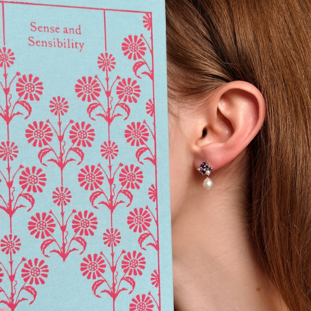 Model wearing silver Beauty Spot amethyst and pearl drop earrings beside a Sense and Sensibility book
