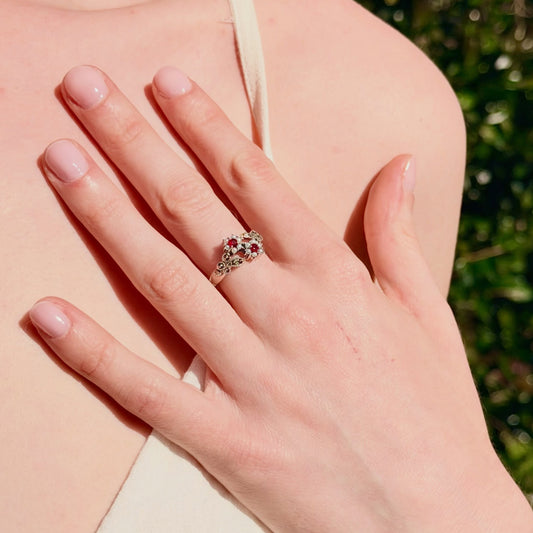 A close-up of a hand wearing a silver-toned ring with small clear stones and a central cluster of red gemstones, shown against skin and a soft green blurred background.